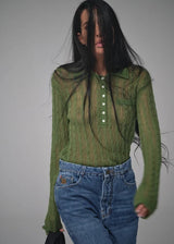 A woman wearing a green ribbed long-sleeve polo shirt with white buttons and blue jeans, photographed against a plain gray studio background.