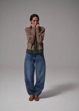 Person wearing a brown sweater, green shirt, blue jeans, and brown shoes on a gray background