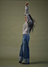 A woman in profile wearing a light gray cable-knit sweater and wide-leg blue jeans with cuffed hems, standing against a solid olive-green studio background.