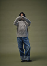 A model wearing a gray cable-knit turtleneck sweater and wide-leg blue jeans with rolled cuffs, standing against a solid olive green studio background.