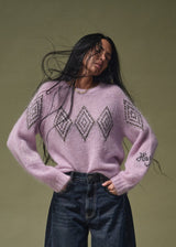 A woman with long dark hair wearing a light pink fuzzy knit sweater with a black diamond pattern and dark blue jeans, posing against a solid olive green background.