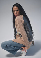 A model with long black hair crouches in a studio setting, wearing a beige cable-knit cardigan over a dark gray quilted jacket, blue jeans, and white sneakers.