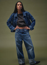 A woman modeling a blue denim jacket and matching wide-leg jeans with rolled cuffs, worn over a dark top, against a solid olive-green studio background.