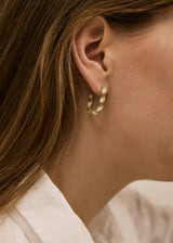 A close-up of a model's ear wearing a small gold hoop earring with a textured, woven or beaded gold-tone design.