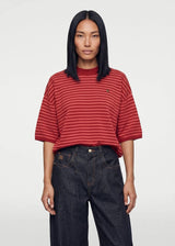 A model wearing a red and dark red striped short-sleeve t-shirt with a small embroidered logo on the left chest, paired with dark blue jeans, against a light gray background.