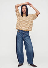 A model wearing a cropped, short-sleeve striped knit top in beige and brown, paired with wide-leg blue jeans and dark brown pointed-toe heels, posing against a light gray studio background.
