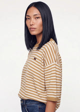 A woman with straight black hair is shown in profile wearing a beige and white horizontally striped short-sleeve t-shirt with a small embroidered logo on the chest, paired with blue jeans, against a plain white background.