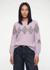 A model with straight black hair wearing a light pink fuzzy knit sweater with a gray geometric diamond pattern across the chest and sleeves, paired with dark blue jeans, against a plain white studio background.