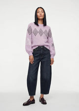 A model stands against a light gray studio background wearing a light purple knit sweater with a geometric diamond pattern on the chest and sleeves, dark wide-leg jeans, and dark brown leather loafers.