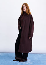 A woman with red hair stands in a studio wearing a long dark brown coat over a striped top and black wide-leg pants, against a white backdrop with a blue floor.
