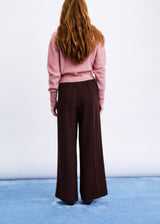 A woman viewed from behind wearing a pink knit sweater and dark brown wide-leg trousers, standing against a white wall on a blue floor.