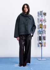 A model stands in a studio wearing a dark green and black checkered cropped jacket with a hood, paired with black wide-leg pants. To the right is a clear acrylic photo display stand against a white wall and light blue floor.