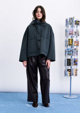 A model stands in a studio wearing a dark green and black houndstooth cropped jacket with a high draped collar, paired with wide-leg black leather trousers. To the right is a clear acrylic display stand filled with postcards against a white wall and light blue floor.