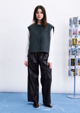 A model stands in a studio setting wearing a sleeveless black and white houndstooth vest over a white long-sleeve shirt, paired with black wide-leg faux-leather pants and dark brown shoes. A white wall and a blue carpeted floor are visible, with a magazine rack on the right.