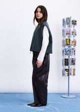 A woman stands in profile wearing a dark green and black houndstooth vest over a white long-sleeve shirt and dark wide-leg trousers. She is standing on a light blue carpet against a white wall, next to a tall display stand filled with small printed photographs.
