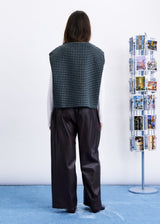 A model stands with their back to the camera wearing a sleeveless black and white gingham vest over a white long-sleeve shirt and dark wide-leg trousers. To the right is a tall, narrow display rack filled with small printed cards against a plain white wall and blue floor.