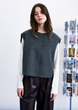 A woman modeling a sleeveless black and white gingham knit top layered over a white long-sleeve shirt and black pants, standing next to a display stand of photographs against a white wall.