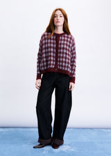 A woman stands against a white studio wall wearing a burgundy and dark red checkered cardigan, loose black wide-leg jeans, and brown shoes.