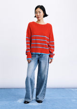 A model stands against a white studio wall wearing a bright red knit sweater with light blue horizontal stripes and wide-leg light-wash blue jeans.