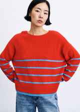 A model wearing a bright red knit sweater with light blue horizontal stripes across the chest and sleeves, paired with blue jeans, against a plain white background.