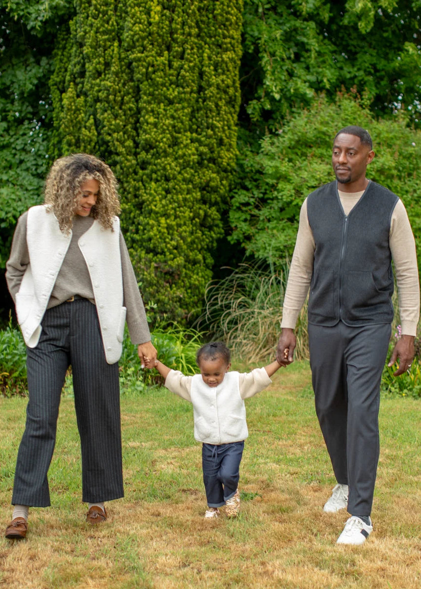 A family of three walking hand-in-hand across a grassy lawn with a large green tree in the background. The woman wears a white sleeveless vest over a taupe top and dark pinstripe trousers.
