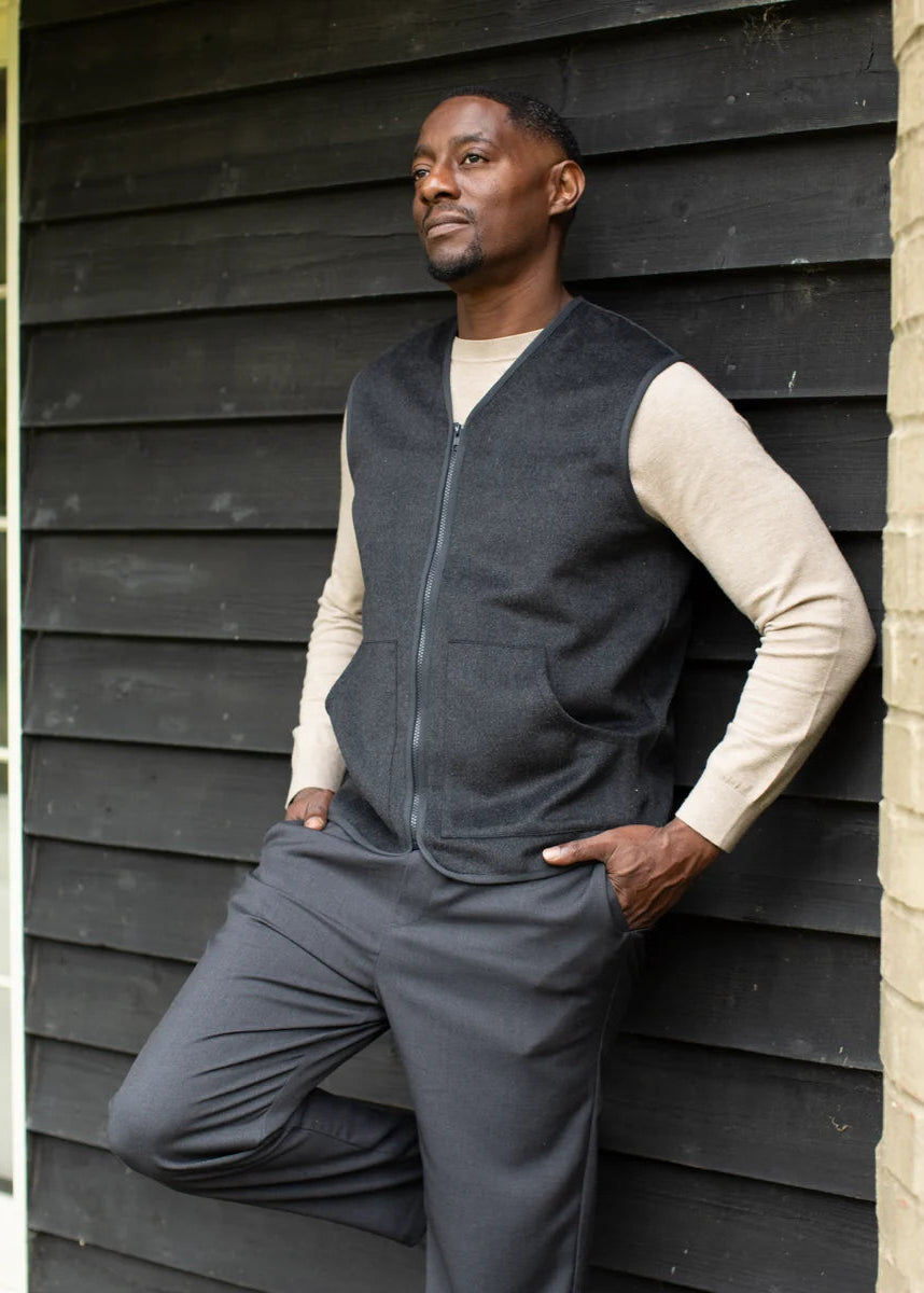 A man wearing a dark gray zippered knit vest over a beige long-sleeve shirt and matching dark gray trousers, leaning against a black wood-paneled wall.
