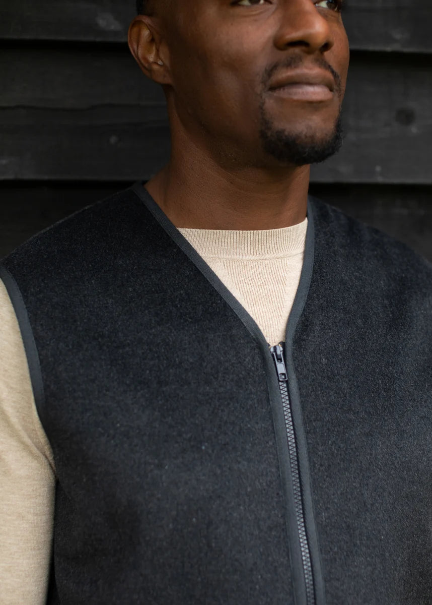 A man wearing a dark charcoal gray zip-up vest over a beige long-sleeve shirt, standing against a dark wooden plank wall.