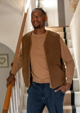 A man wearing a brown fleece vest over a tan crewneck sweater and navy drawstring trousers, standing on a staircase indoors.
