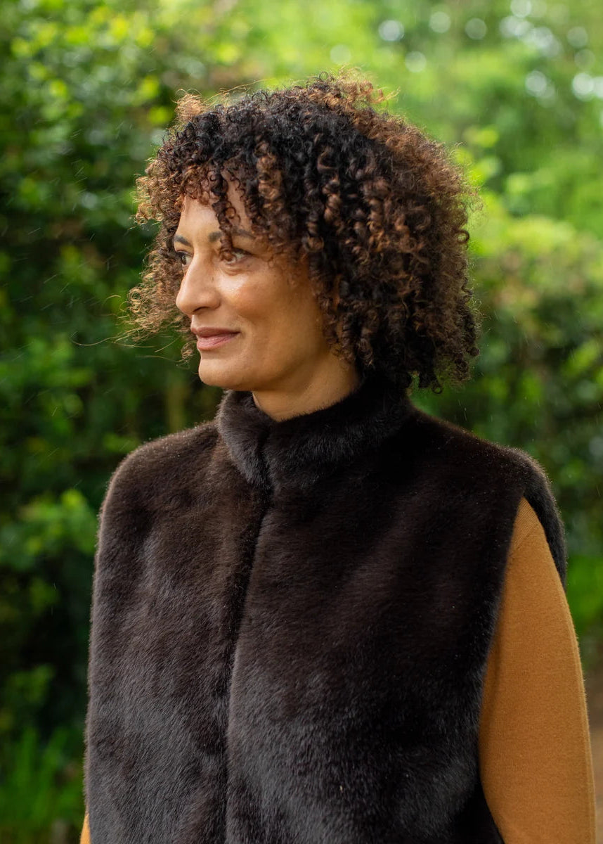 A woman with curly brown hair wearing a dark brown fur vest over a tan top, standing outdoors against a blurred green foliage background.