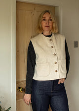 A woman wearing a cream-colored wool vest with brown buttons over a dark gray long-sleeve top and dark blue jeans, standing in front of a light wood door.