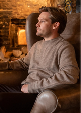 A man in a warm brown armchair wearing a taupe knit sweater, holding a pint of beer in a cozy, dimly lit pub setting with a stone fireplace in the background.