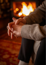 A close-up lifestyle shot of two people sitting together with their hands clasped, wearing ribbed knit sweaters in beige and gray, with a warm glowing fireplace in the background.