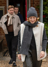 A woman walking outdoors wearing a cream-colored textured vest over a dark gray sweater, paired with a matching dark gray scarf and beanie. Two men are visible in the background near a brick building entrance.