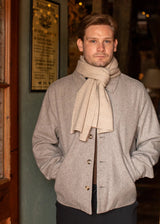 A man wearing a light gray wool-blend jacket and a beige knit scarf, standing in a doorway of a rustic shop.