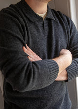 A man wearing a dark charcoal gray knit polo sweater over a light beige undershirt, with his arms crossed.