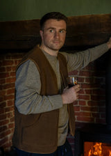 A man wearing a brown wool vest over a gray knit sweater stands in front of a brick fireplace, holding a glass of red wine.