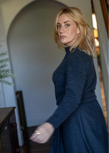 A woman with blonde hair wearing a dark navy blue long-sleeve mock-neck dress, standing in an indoor setting with a blurred arched doorway in the background.