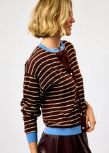 A woman wearing a brown and beige striped cardigan with light blue ribbed trim, shown from a side-back angle against a plain light gray studio background.