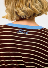 Close-up back view of a person wearing a brown and cream striped long-sleeve shirt with a light blue ribbed crew neck. The brand name 'Shinoko' is embroidered in light blue script on the upper back.