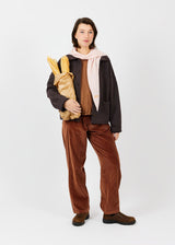 A model stands against a light gray studio background wearing a dark brown jacket, brown corduroy pants, a light pink scarf, and brown boots, while holding a loaf of bread wrapped in paper.