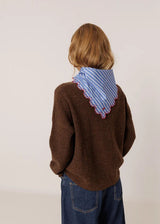 A child viewed from behind wearing a brown knit sweater, blue jeans, and a blue-and-white striped bandana with a scalloped edge around the neck, against a plain light background.