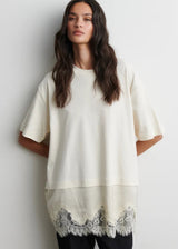 A woman modeling an oversized cream-colored short-sleeve top with a scalloped lace hem, paired with black pants, against a plain light gray wall.