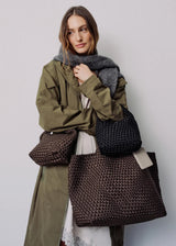 A woman wearing an olive green coat and gray scarf models three woven tote bags in brown, black, and dark brown against a light gray studio background.
