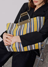 A woman in a black blazer and trousers holds a large striped tote bag with yellow, blue, and white vertical stripes and metallic handles.