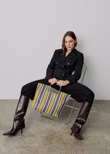 A model sits on a metal chair in a minimalist studio setting, wearing a black double-breasted blazer and matching trousers, dark brown croc-embossed knee-high boots, and holding a striped tote bag with yellow, gray, and blue stripes.
