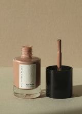 A bottle of nail polish with its cap off and brush visible, set on a beige surface against a matching beige background. The polish color is a muted dusty pink, and the bottle has a white label reading 'WITNESSED PENNY Nail Polish'.