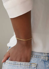 A close-up of a person wearing a delicate gold chain bracelet on their wrist, paired with a white top and light blue denim jeans.