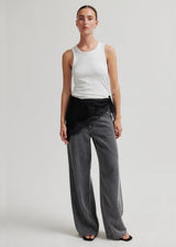 A model stands against a light gray studio background wearing a white ribbed tank top, wide-leg gray jeans with a black draped waist detail, and black sandals.