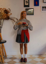 A woman stands in a room with a white textured wall and checkered floor, wearing a black and white striped button-up shirt, a dark gray pleated skirt, white socks, and dark red loafers. She has a bright red fabric sash tied around her waist and is holding a small red cup and saucer.