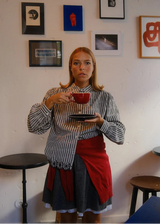 A woman stands indoors holding a red mug and saucer, wearing a black-and-white gingham shirt, a gray skirt, and a red apron tied around her waist. Framed wall art hangs on a white wall behind her, with small black and wooden tables at the sides.